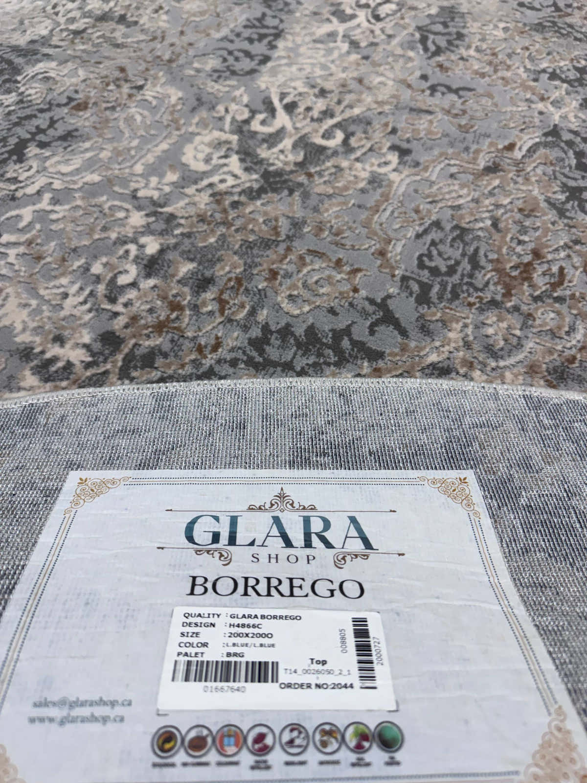 A close-up of a patterned area rug in gray, beige, and brown tones, with a white product tag reading 'GLARA SHOP BORREGO' placed on the foreground edge.