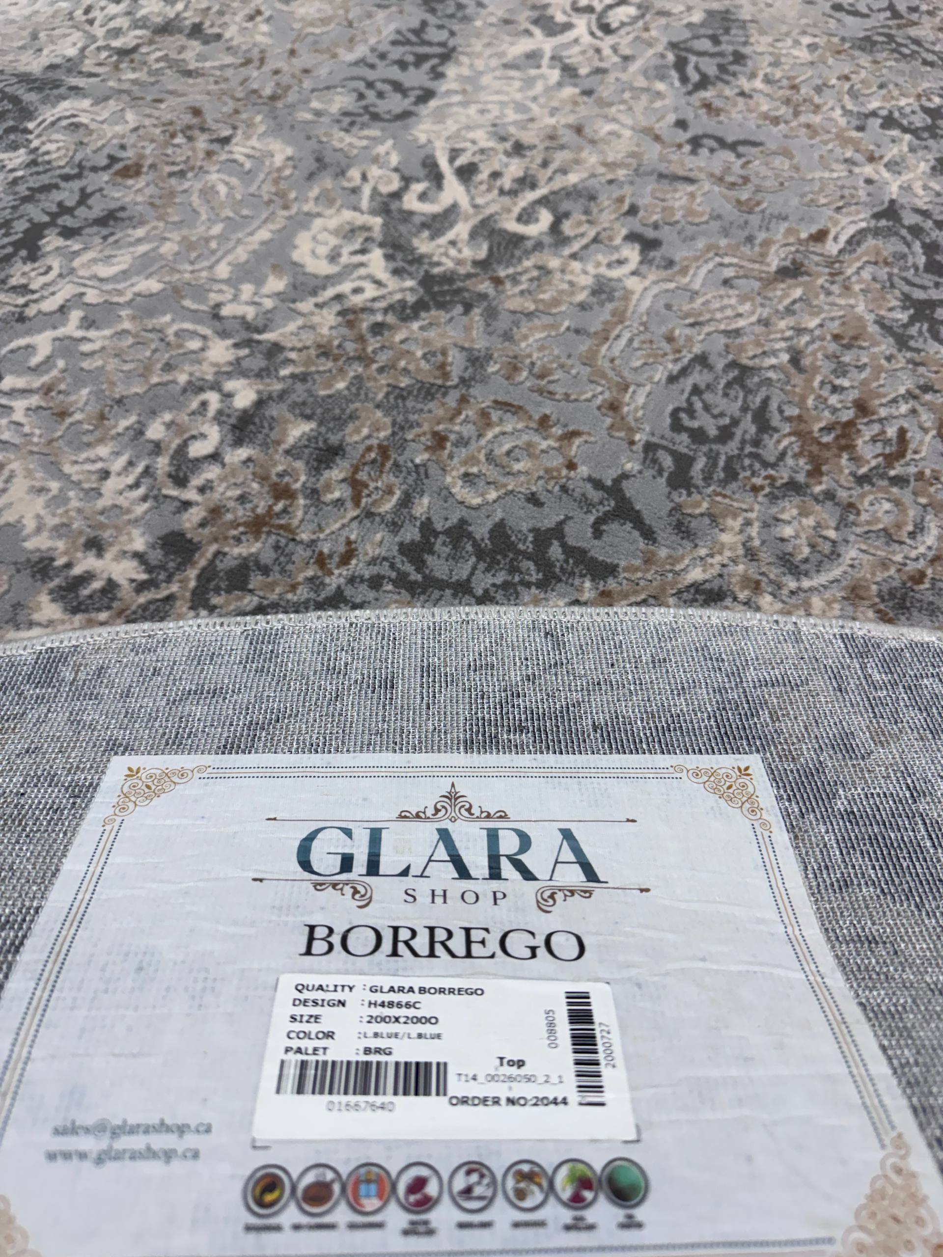 A close-up of a patterned area rug in gray, beige, and brown tones, with a white product tag reading 'GLARA SHOP BORREGO' placed on the foreground edge.