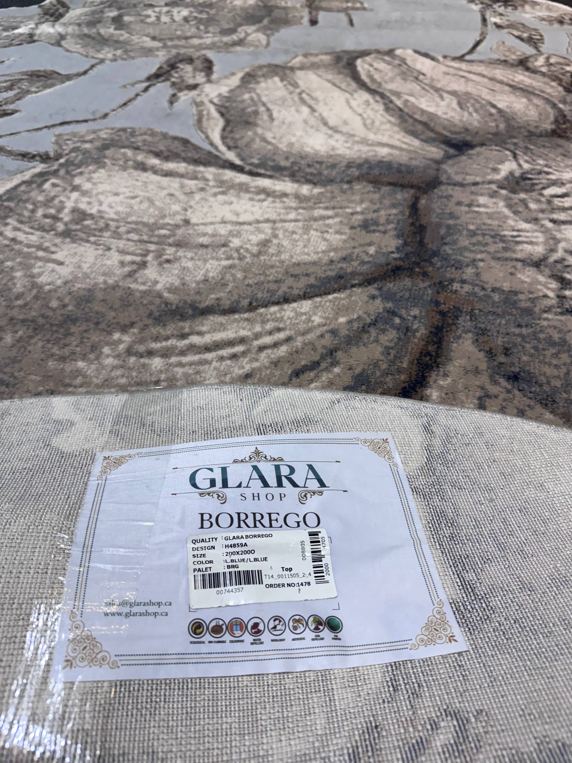 A close-up of a textured woven fabric or rug sample labeled 'GLARA SHOP BORREGO' with a white product tag, placed against a larger matching patterned textile in a gray and beige abstract design.