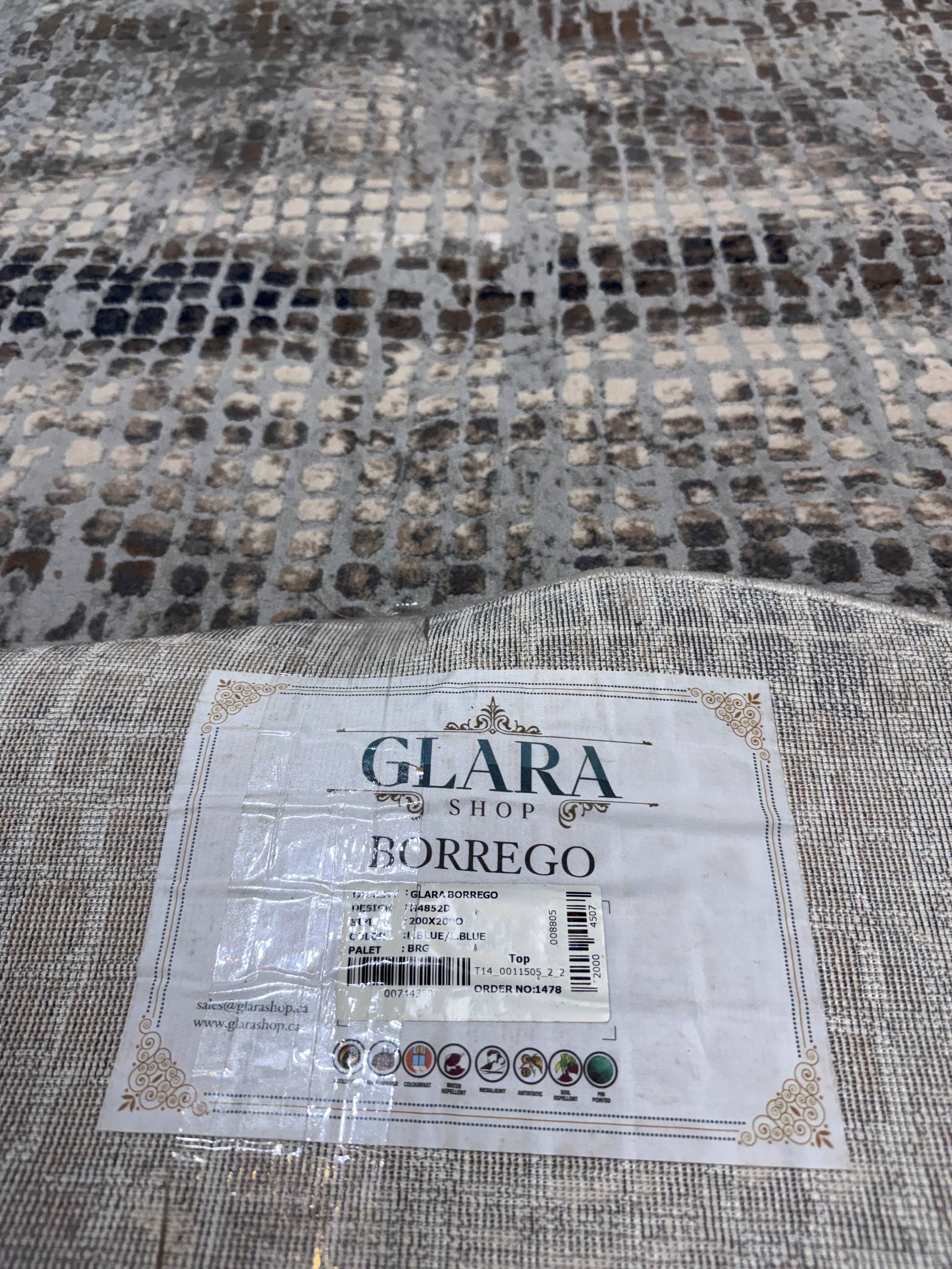 A close-up of a woven rug or textile with a snakeskin-like pattern in beige, gray, brown, and black, with a white paper label reading GLARA SHOP BORREGO attached in the foreground.