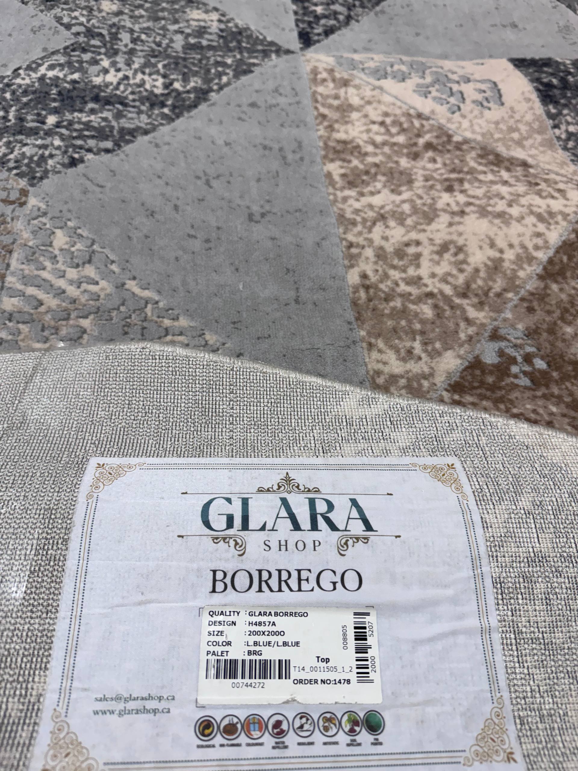 A close-up of a folded rug or textile with a visible care label reading GLARA SHOP BORREGO, placed over a larger patterned rug in gray, beige, and brown tones.