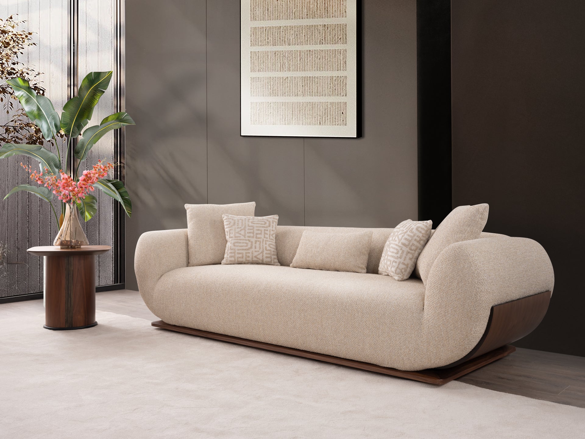 A modern curved upholstered sofa in warm beige fabric with dark wood side panels and base, styled in a minimalist interior with matching pillows, a side table with flowers, and dark gray walls.