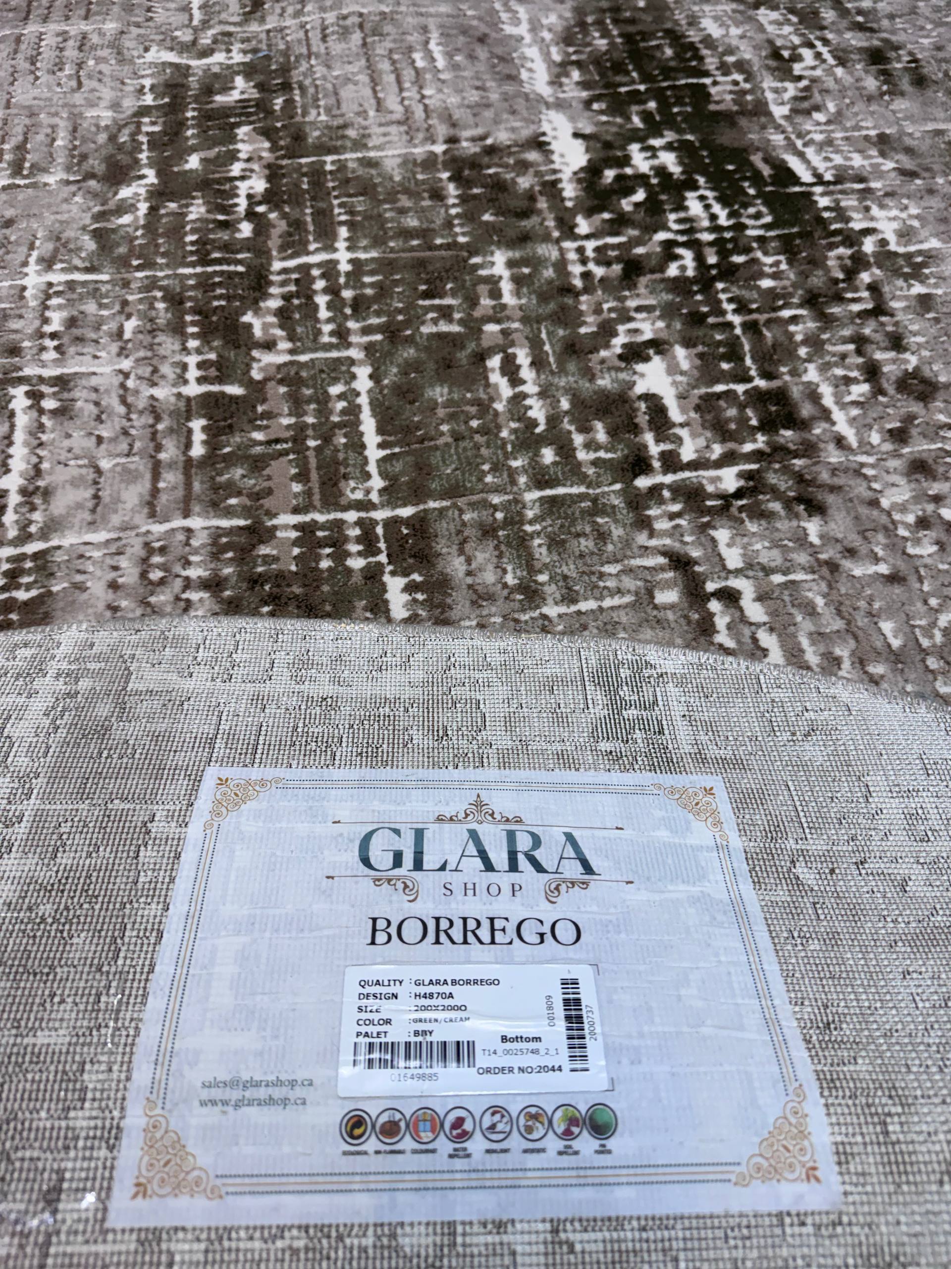 A close-up of a textured area rug with a distressed, abstract pattern in shades of gray, beige, and brown. A white paper label from 'GLARA SHOP' is placed on the rug, identifying the product as 'BORREGO'.